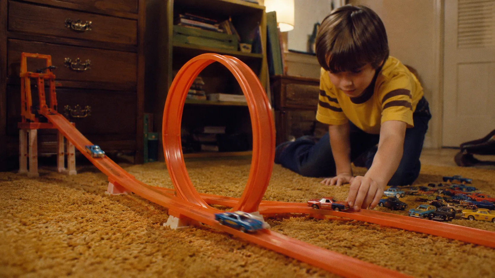 A child racing die-cast cars through a bright orange Hot Wheels loop on 1970s shag carpet