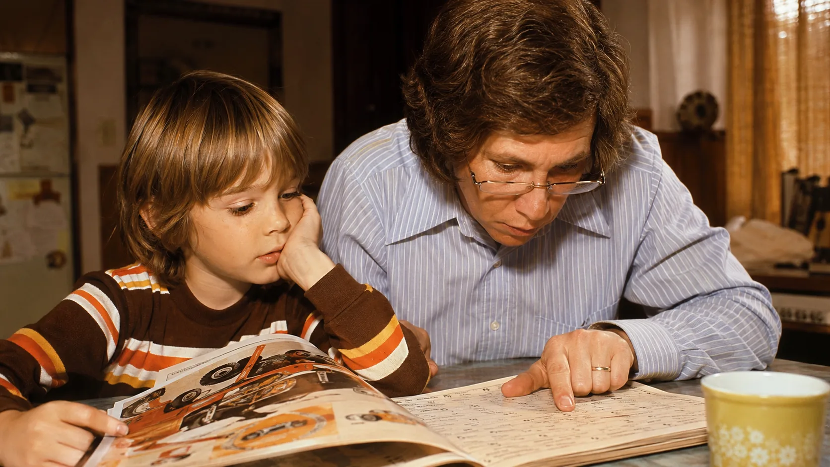 A father wearing reading glasses studying the fine print in a 1970s toy catalog with his child