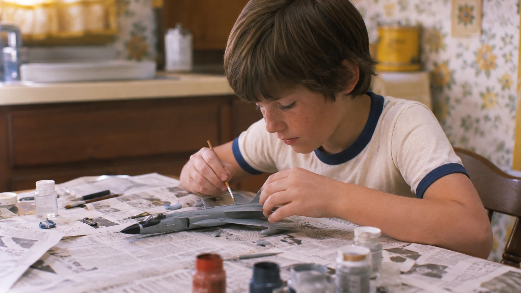 Child carefully painting a plastic model airplane at a newspaper-covered 1970s kitchen table