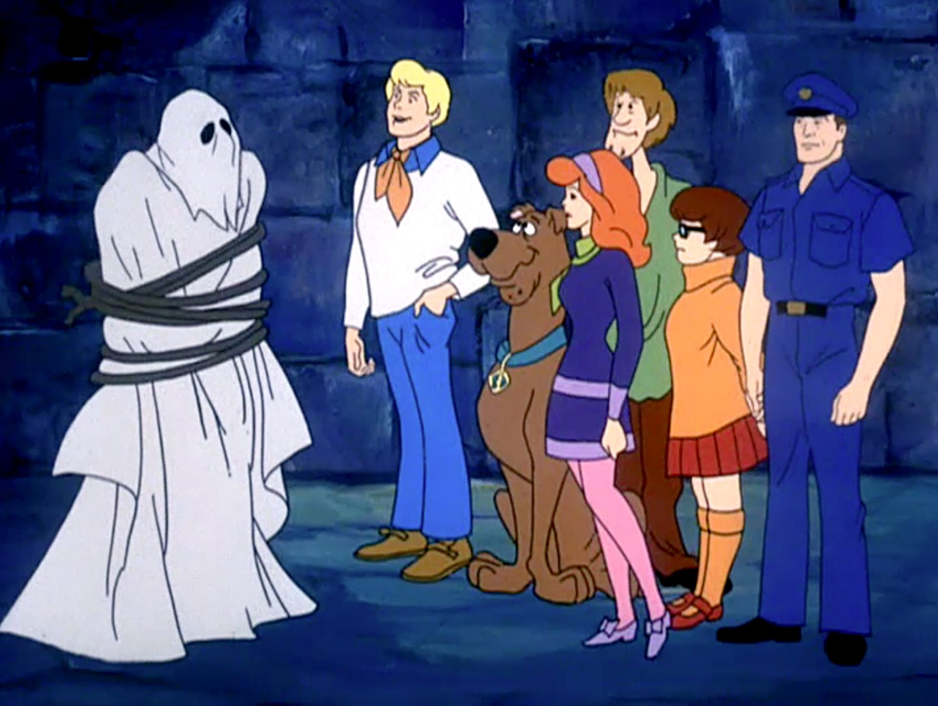 Fred, Daphne, Velma, Shaggy and Scooby-Doo standing beside an unmasked ghost and a police officer