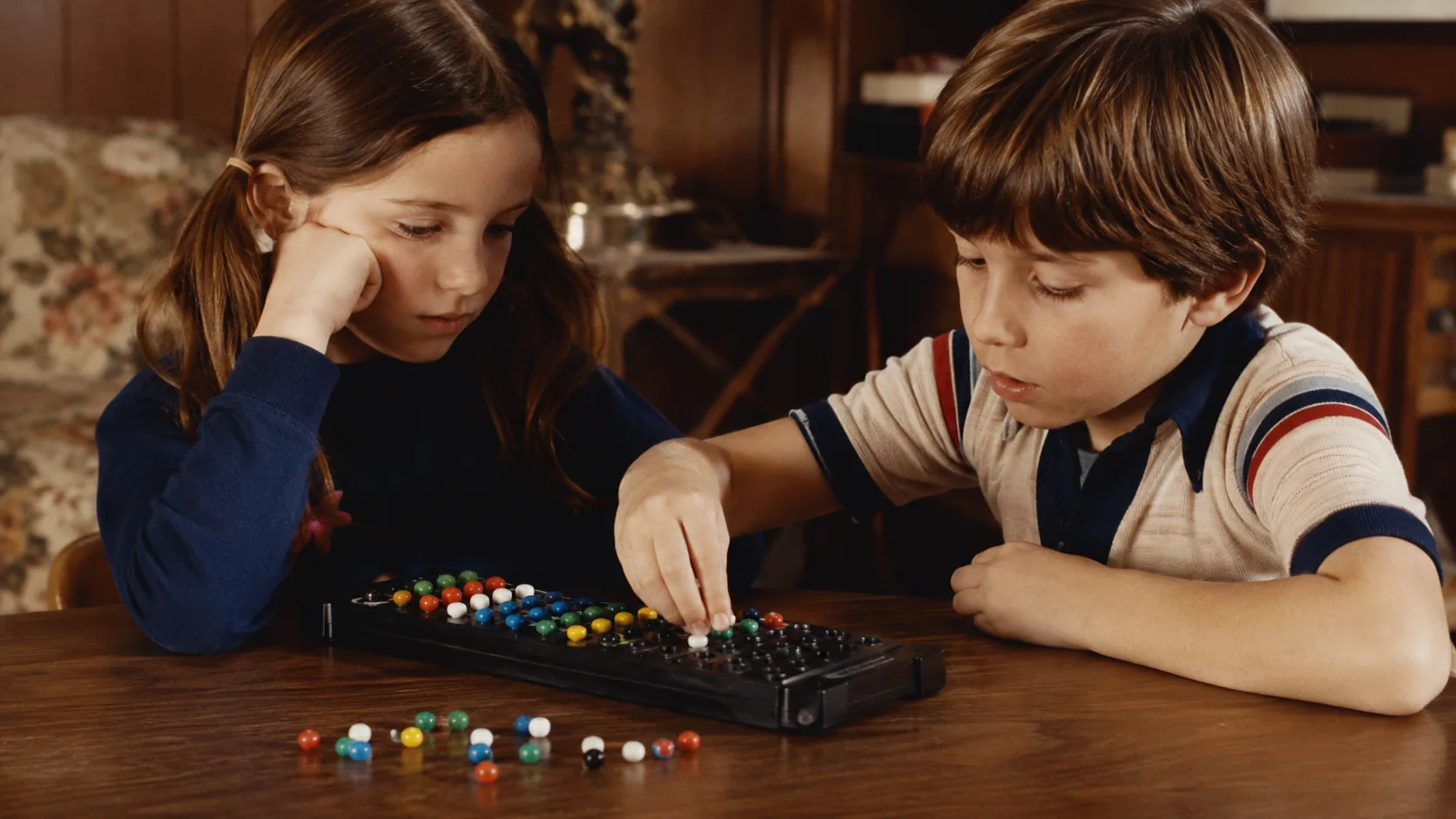 Two children concentrating on a colorful code-breaking board game in a 1970s family room