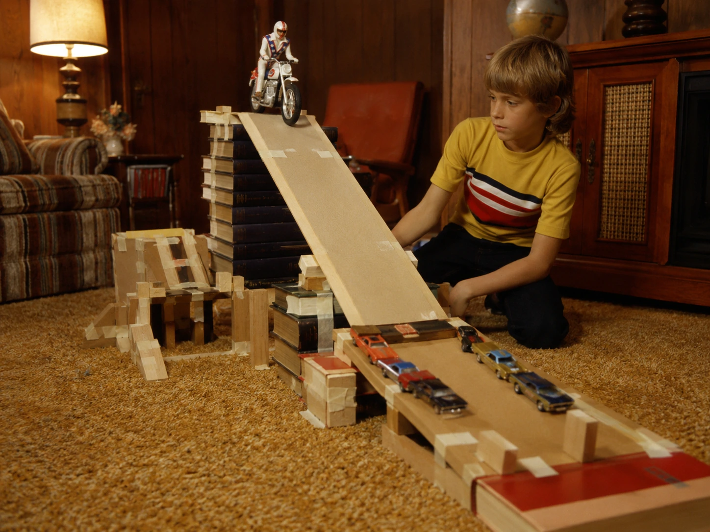 Child building a large Evel Knievel ramp from books, cardboard, wooden blocks and tape