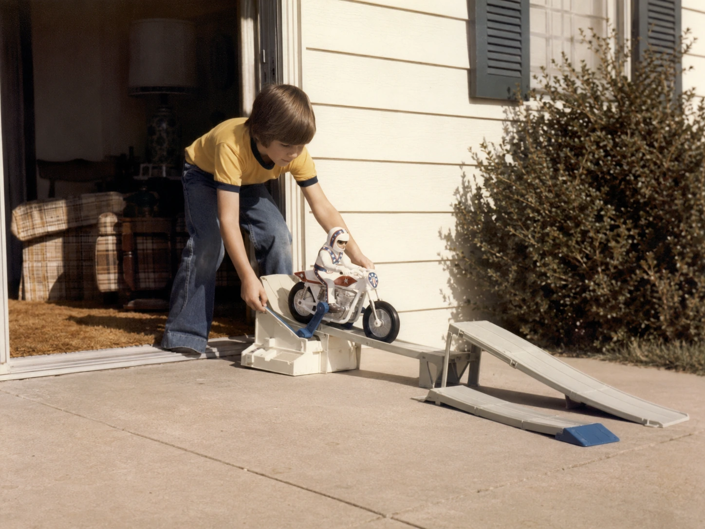 Child moving an Evel Knievel Stunt Cycle setup from a family room onto a sunny driveway