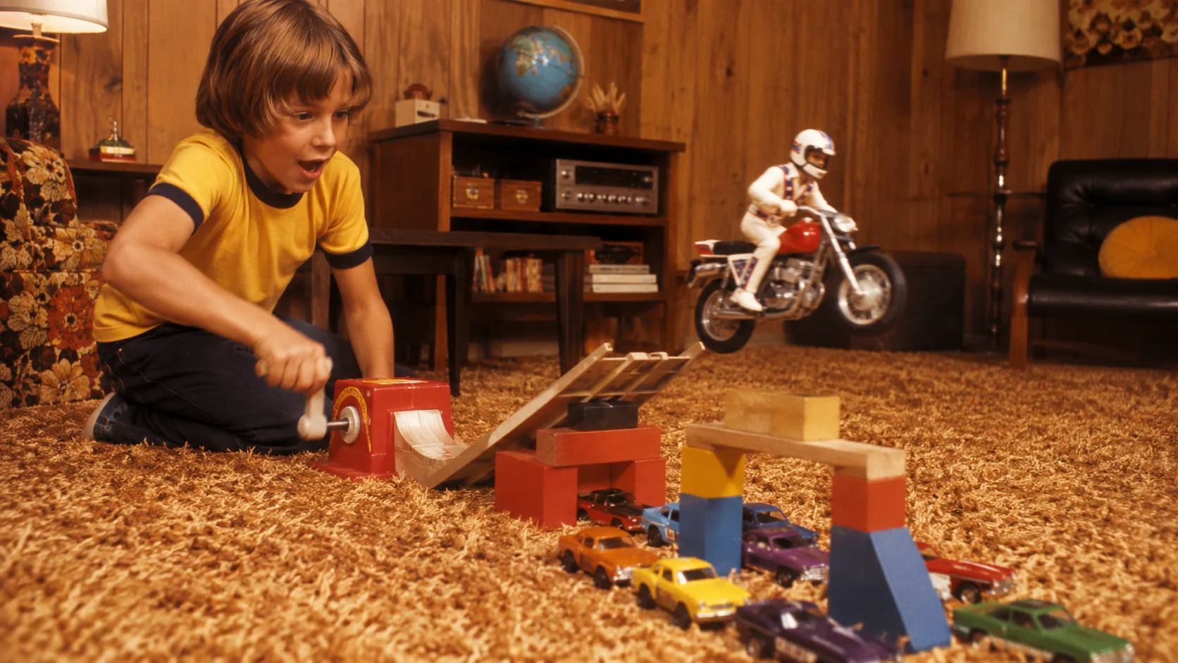 Child launching an Evel Knievel stunt motorcycle from a crank-powered ramp over toy cars and blocks