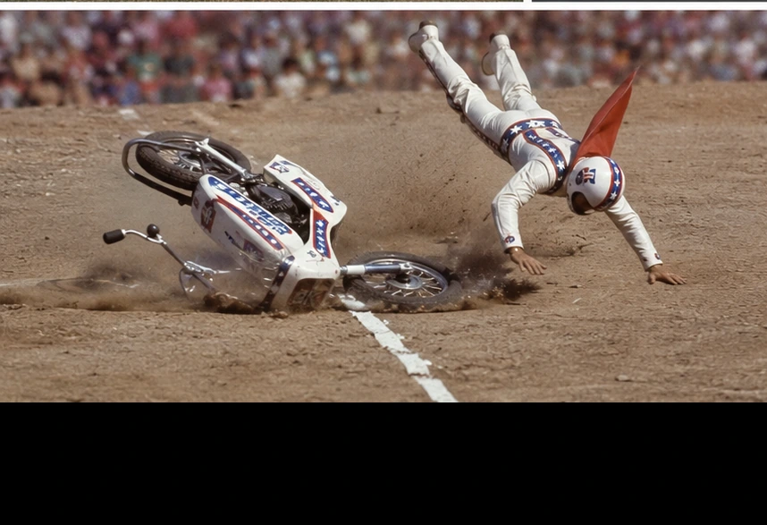 Evel Knievel toy figure thrown from a motorcycle beside a red ramp on dirt