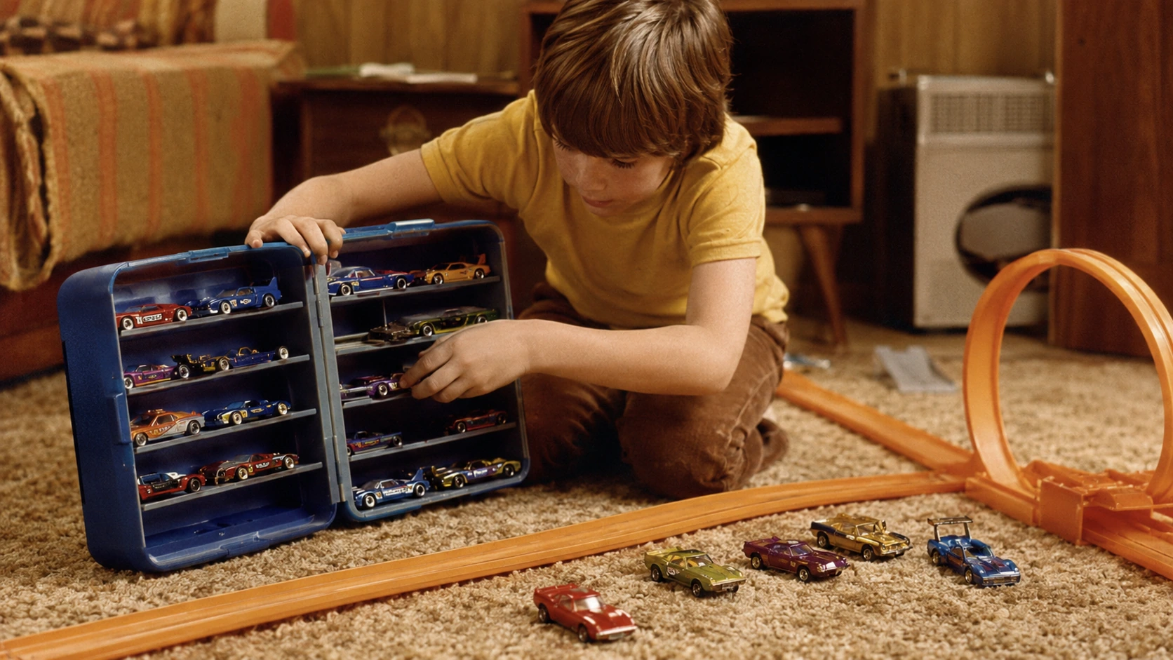 A child opening a blue carrying case filled with early custom-style Hot Wheels cars beside orange track in a 1970s playroom