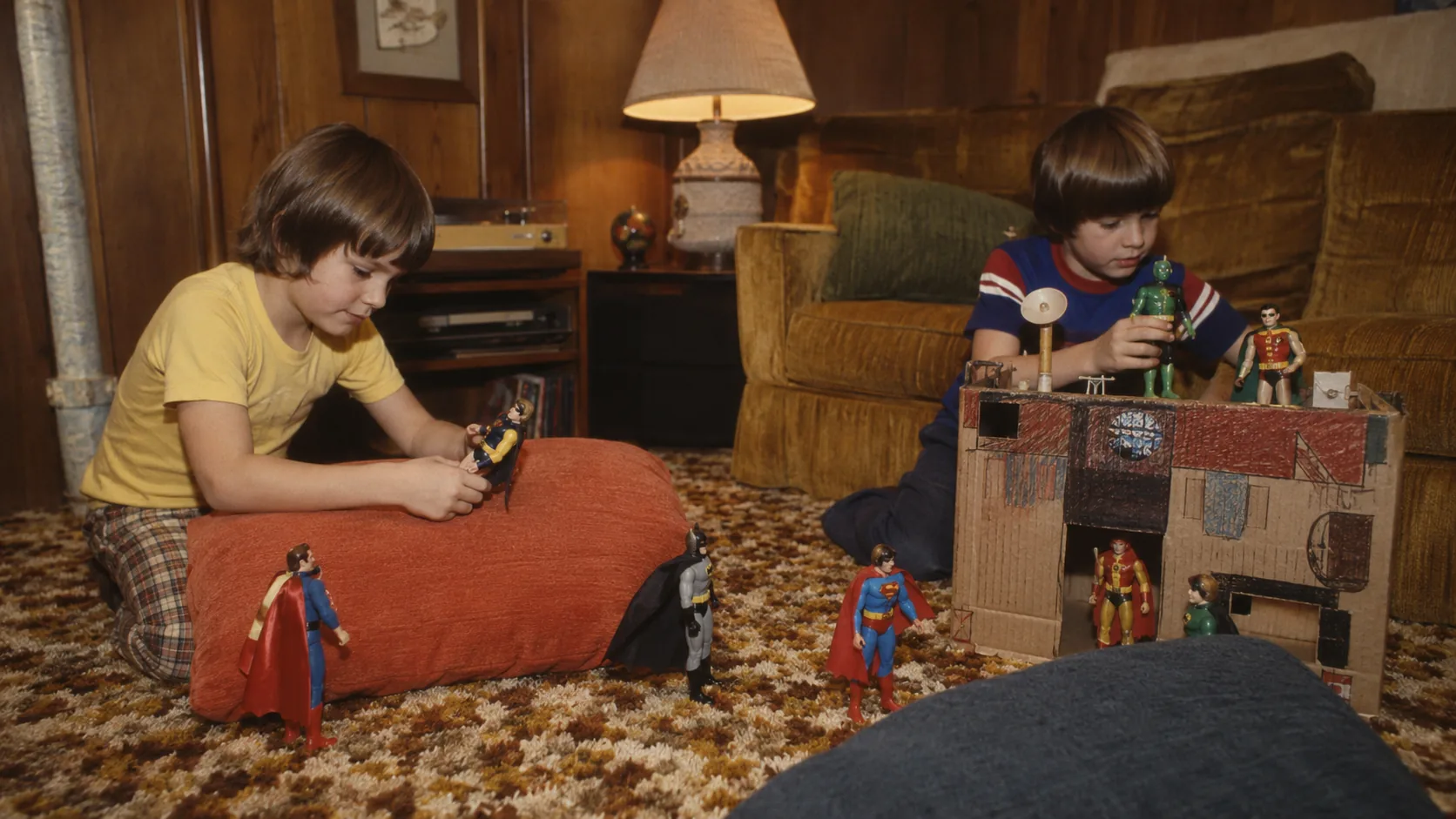 Children staging a 1970s living-room battle with cloth-costumed Mego-style superheroes and a homemade cardboard headquarters