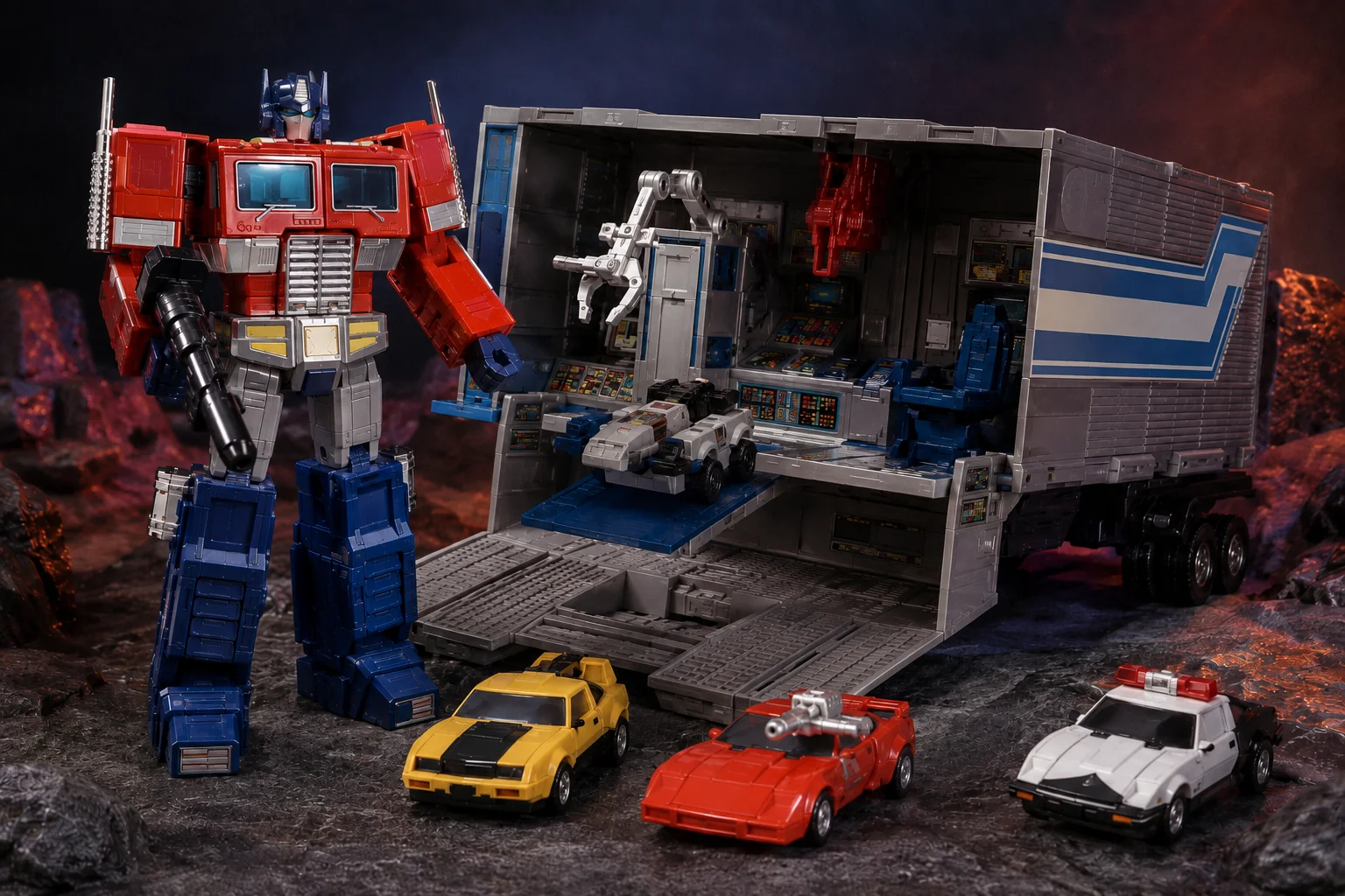 Red-and-blue semi-truck robot posed beside an opened gray command trailer with scout vehicle, repair arm, weapons, and smaller heroic car robots.