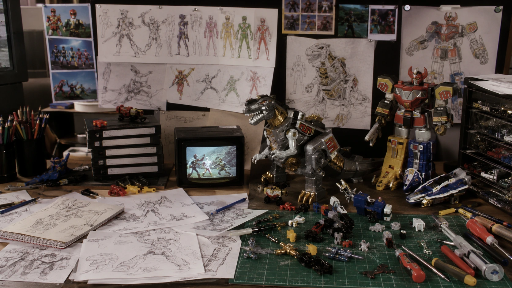 1990s toy development workbench with Ranger-style sketches, dinosaur robot prototypes, VHS tapes and transforming toy parts