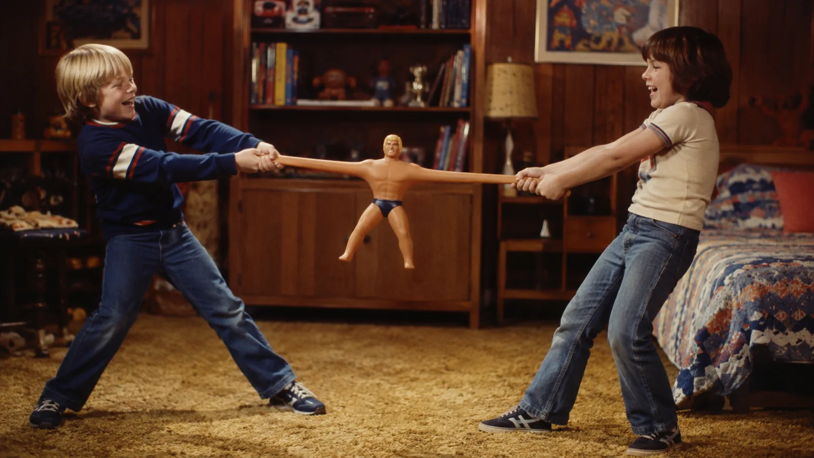 Two children pulling a Stretch Armstrong figure across a 1970s bedroom floor